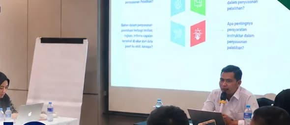 Pelatih memaparkan materi di depan layar presentasi